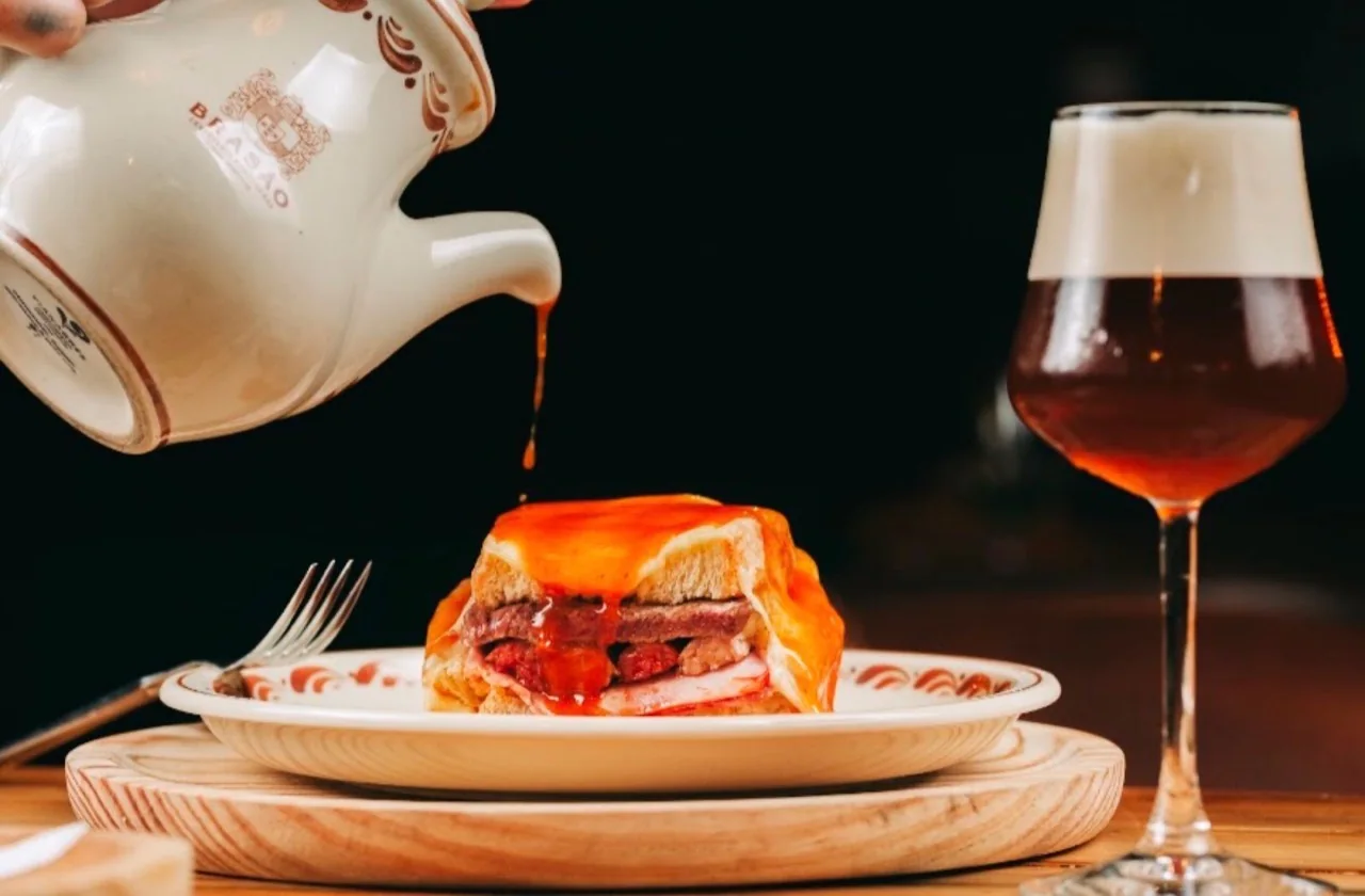 Francesinha at Brasão Aliados as its sauce is poured, with a dark beer beside it.