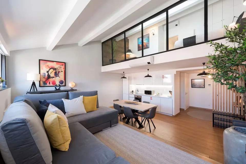 Living and dining area beneath a glazed mezzanine, with a grey sofa and white kitchen in the Batalha duplex.